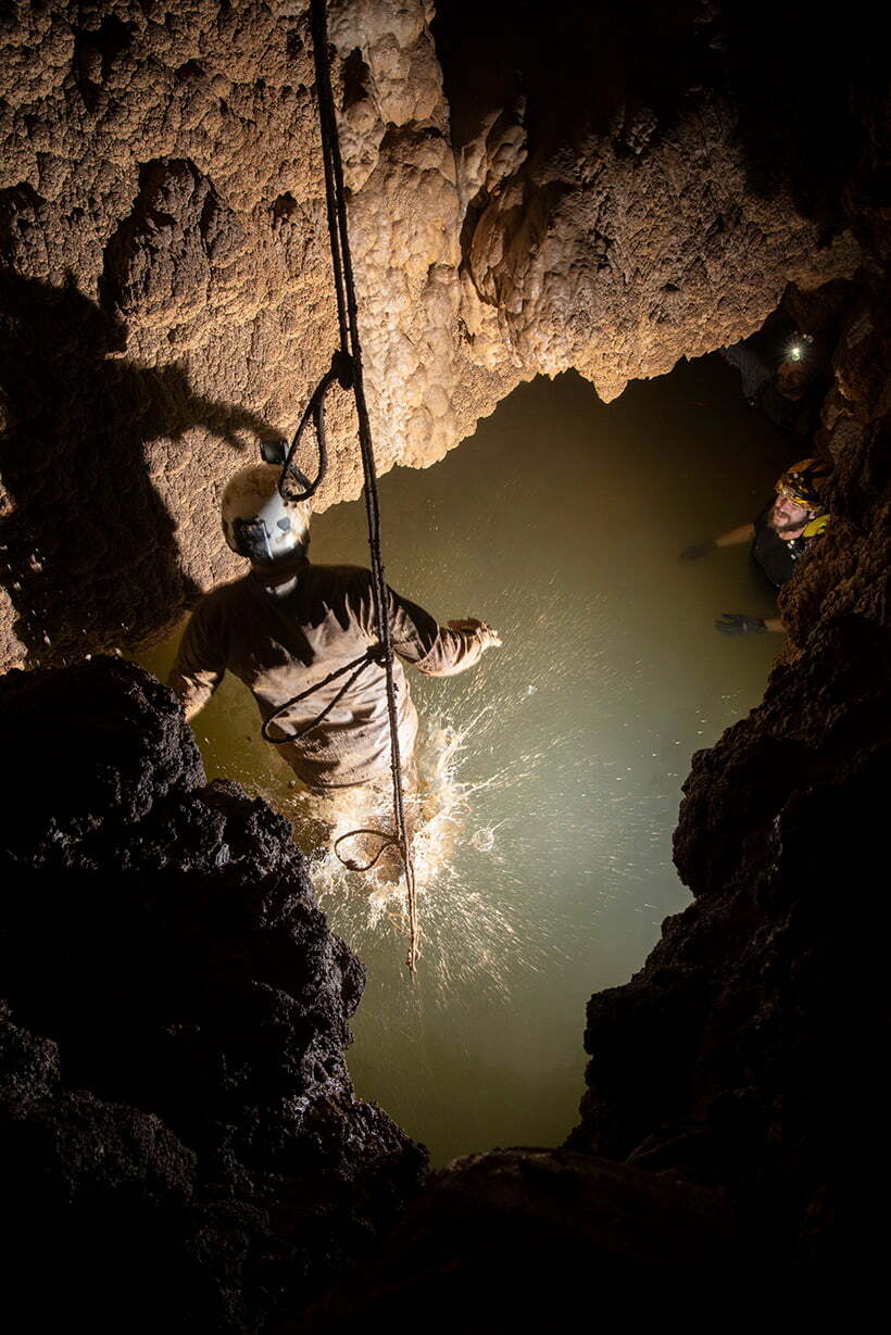 Travis Wuest drops into a passage filled with water in Wild Cave, expedition members Nathan Hinkie and Brad Wuest wait in the background.